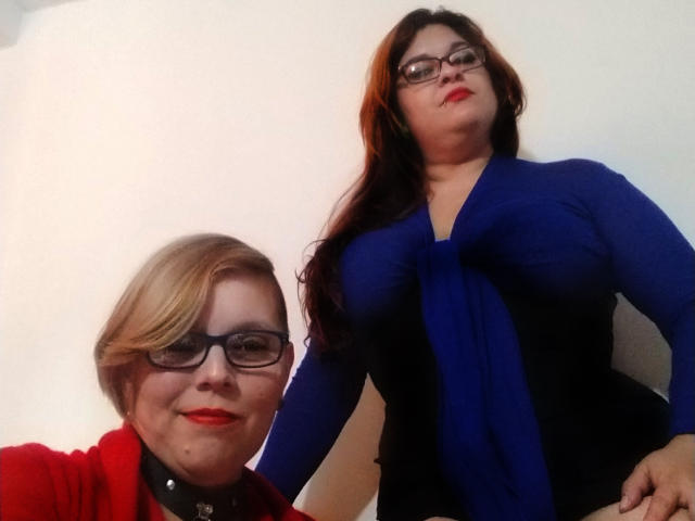 Dos chicas latinas con gafas; una con cabello rubio corto, lápiz labial rojo, blusa roja y cuello negro, la otra con cabello castaño largo, blusa azul y lápiz labial rojo..