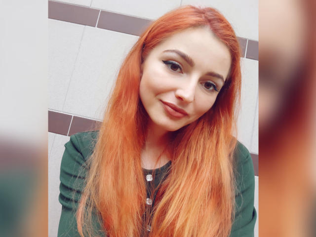 KellyMareta mit langen roten Haaren, grünen Augen und einem leichten Lächeln, die ein grünes Hemd trägt, hat ein süßes Gesicht und eine kleine Nase und blickt direkt in die Kamera..