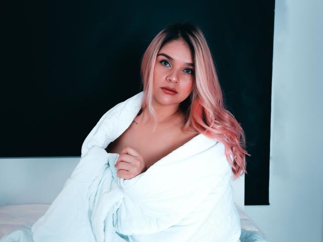 ValerieStone, avec ses cheveux roses, ses yeux bleus et sa peau pâle, partiellement couverte par une couverture blanche, exposant ses seins, assise sur un lit, regardant directement l'appareil photo..