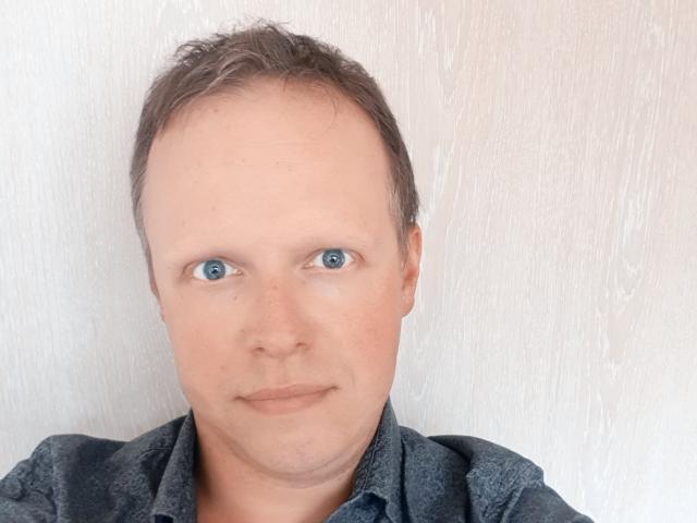 NaughtyHugues a une tête dégarnie avec des cheveux bruns fins, des yeux bleus et un visage légèrement rond, et porte une chemise bleu foncé à motifs..