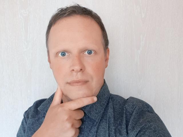 NaughtyHugues has blue eyes, a balding head, and is touching his chin with a finger. He's wearing a dark blue, textured shirt.