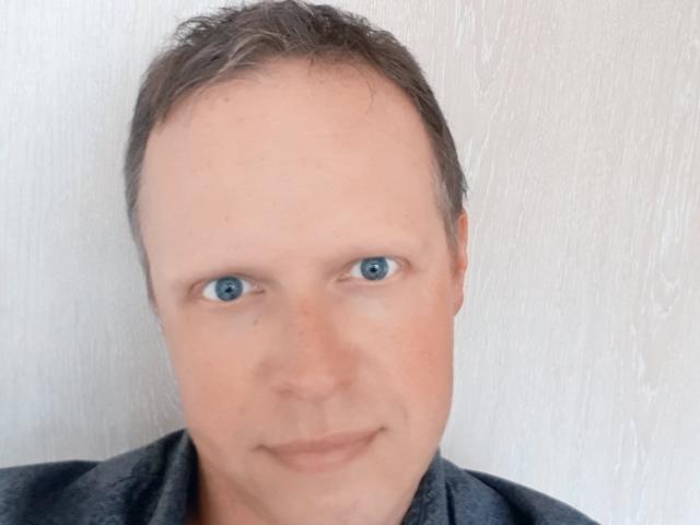 NaughtyHugues a une tête dégarnie avec des cheveux bruns, des yeux bleus et un léger sourire. Il porte une chemise sombre, avec un fond clair et texturé..