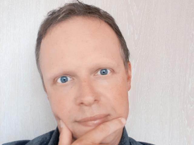 Hugues, travieso, con ojos azules, pelo castaño corto y una expresión neutra, apoyando el mentón en la mano, vistiendo una camisa oscura. Su rostro está ligeramente calvo..