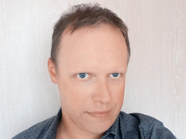NaughtyHugues tiene la cabeza calva con poco cabello castaño, ojos azules y una expresión neutra. Lleva una camisa gris oscuro, frente a una pared blanca con textura..
