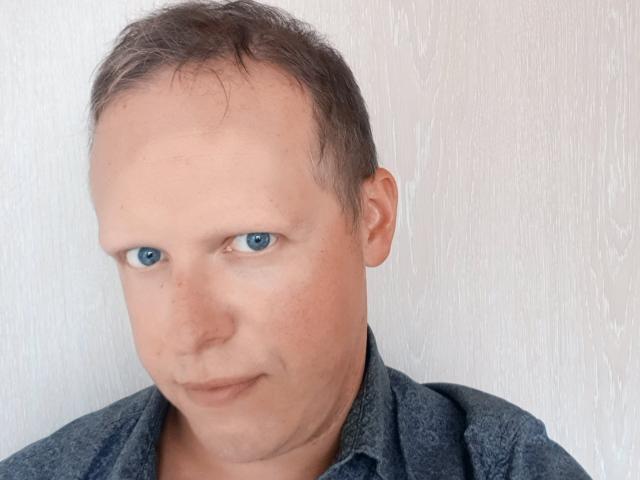 NaughtyHugues tiene la cabeza calva, ojos azules y una ligera sonrisa, y lleva una camisa gris oscuro. Su piel pálida tiene algunas pecas. Sus glúteos no son visibles..