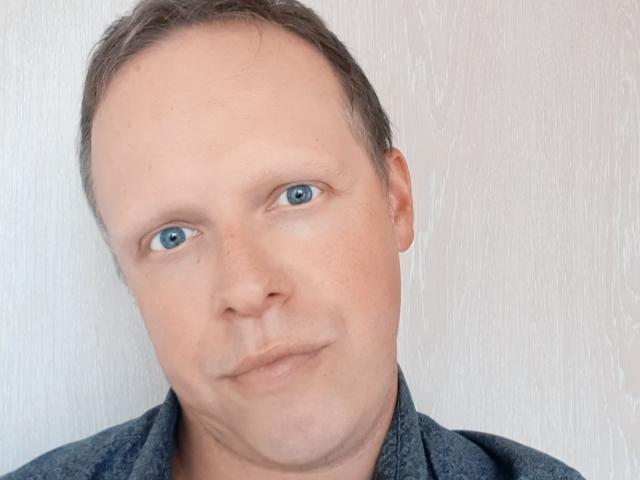 NaughtyHugues, ein Mann mittleren Alters mit heller Haut, blauen Augen und einem leicht verrückten Lächeln, der ein graues Hemd trägt, kurze braune Haare hat und ein paar Sommersprossen..