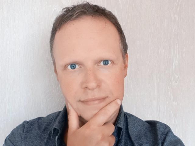 NaughtyHugues met blauwe ogen, kort bruin haar en een lichte glimlach, gekleed in een donker shirt, die zijn kin aanraakt met een hand. Witte, gestructureerde achtergrond..
