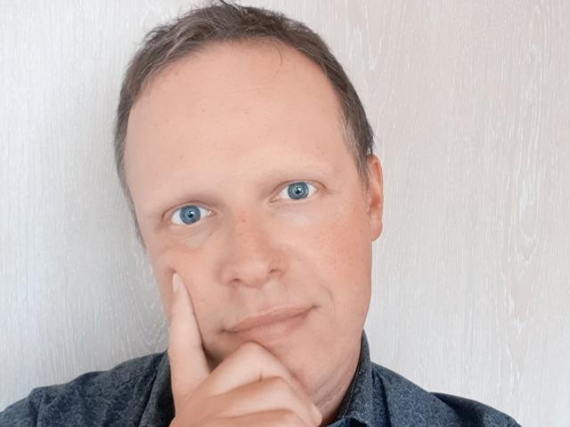 NaughtyHugues hat einen kahl werdenden Kopf mit kurzen, braunen Haaren, blaue Augen und ein leichtes Lächeln, wobei er mit seinem Finger sein Kinn berührt und ein dunkel gefärbtes, strukturiertes Hemd trägt..