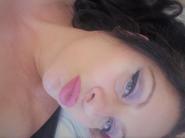 Emerald's face, lying down, with dark wavy hair, pink lipstick, and blue eyeshadow. Her eyes are open, and she's looking at the camera with a slight pout.