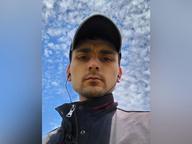 DylanNeasq, con un'espressione seria, indossa un berretto nero e delle cuffie, guarda in basso, con una giacca grigia e nera, sullo sfondo di un cielo blu con nuvole bianche sparse..