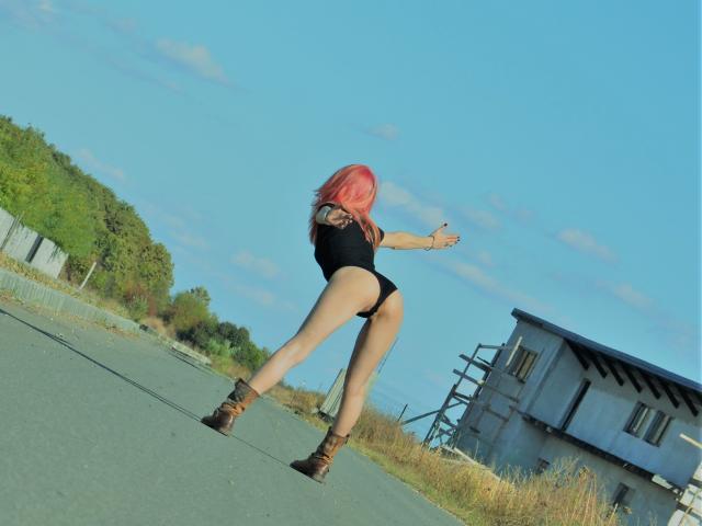 MissCelestine with pink hair, black one-piece, and brown boots, facing away, pointing, showing her ass, on a road, near a blue sky and a gray building.