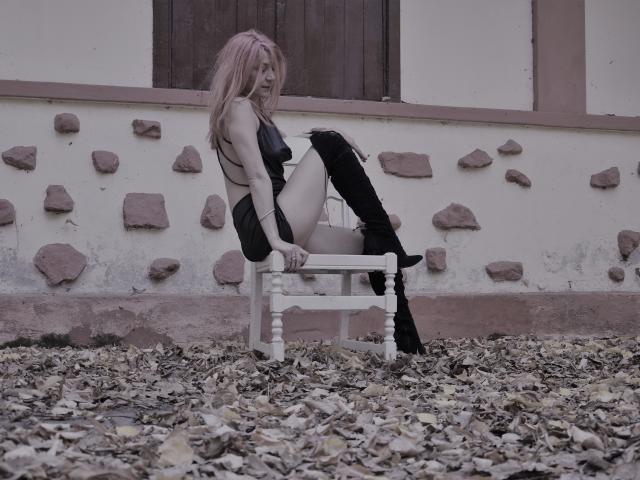 MissCelestine with pink hair sits on a white chair, wearing black lingerie and thigh-high boots, showing her ass and small tits, on a leaf-covered ground.