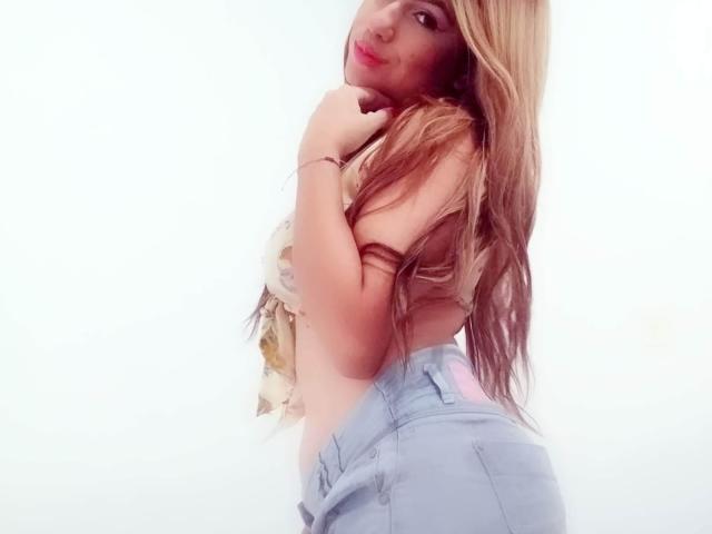 AnastaciaMonaco with long blonde hair, topless, showing her tits, wearing light blue shorts, hand on neck, looking at camera, smiling. Soft light, white background.