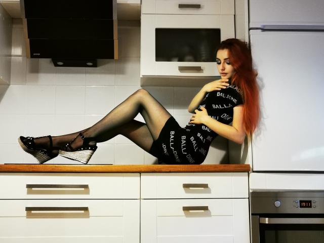 KellyMareta mit roten Haaren, einem schwarzen "Balenciaga"-Kleid, schwarzen Strumpfhosen und schwarzen High Heels, liegt auf der Küchenarbeitsplatte, die Beine angezogen, wobei ihr Hintern zu sehen ist, die Hände auf der Brust, und blickt nach unten..