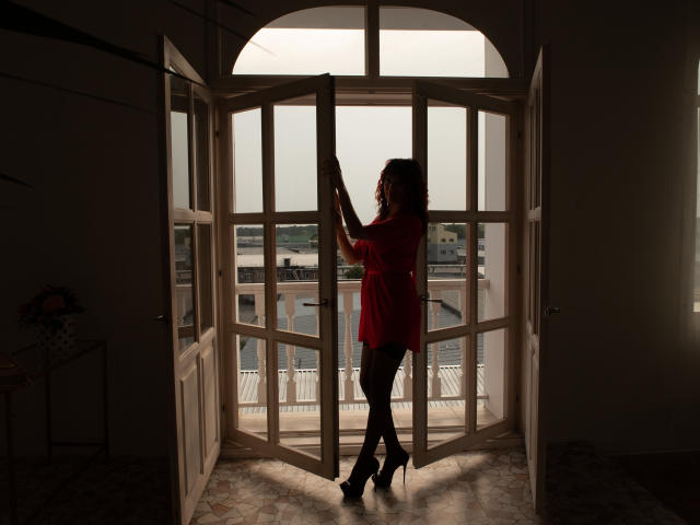 JennySienna staat in een deurpost in het zonlicht, ze draagt een rood shirt, zwarte broek en hoge hakken, en haar silhouet is te zien tegen een raam. Ze staat met haar rug naar de camera, haar handen op het kozijn..
