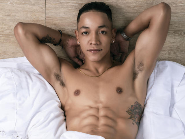 MathewDell, shirtless, lying on white bed, arms behind head, brown skin, toned abs, small nipples, armpit hair, tattoo on right side, short black hair, gold necklace, bracelet.