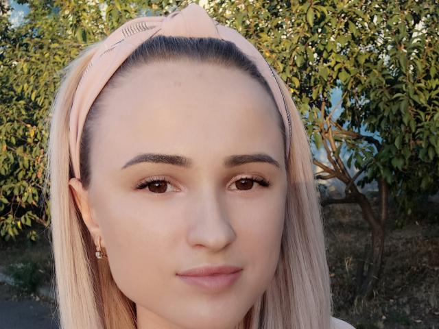 EllenXParadise has pale skin, brown eyes, and straight blonde hair with a pink headband. She's smiling subtly with a small nose and wearing small gold earrings.