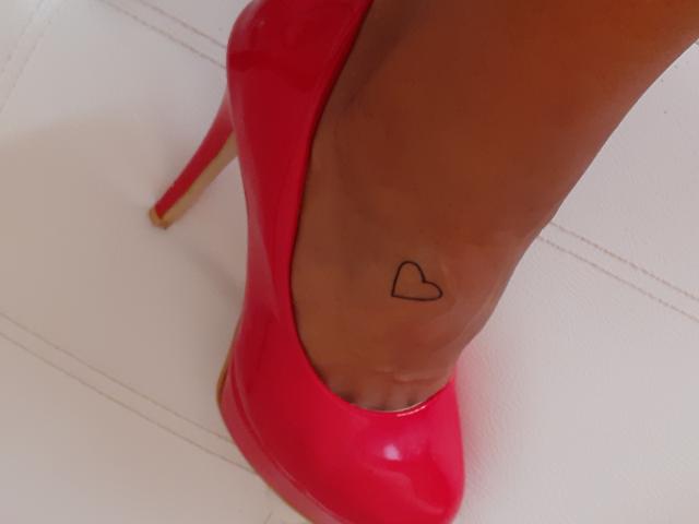 CassandraMichelli's foot in a shiny red high heel, showing a small black heart tattoo on her ankle. Her skin is dark brown. The heel is glossy and pointed.