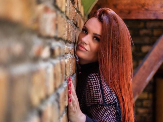 RahelHot with long red hair, wearing a black mesh top, leaning against a brick wall, looking over her shoulder with a slight smile, red nails.