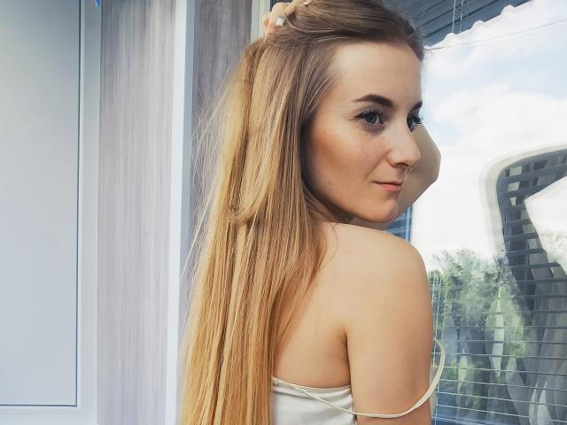ValerieStone, seminua, deitada em uma cama, vestindo shorts jeans desgastados, olhando para a câmera, com cabelo rosa, olhos escuros e uma expressão sexy e confiante. Suas nádegas estão elevadas..
