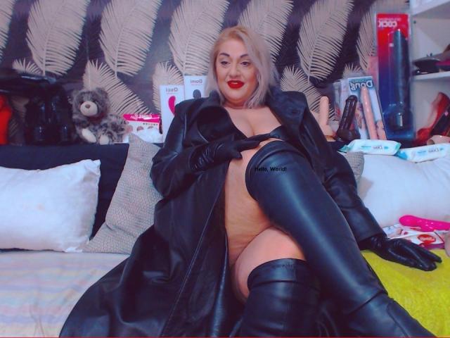 SexyyMilf with blonde hair, wearing a black leather skirt, red thigh-high boots, and red gloves, on all fours, showing her ass and thick thighs, in a bedroom.
