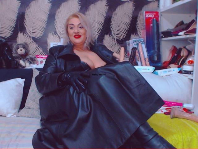 SexyyMilf with blonde hair, purple lipstick, and red gloves, wearing a black leather corset, showing her big boobs, in a bedroom with feather-patterned wallpaper.