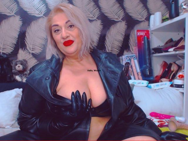 SexyyMilf, blonde avec du rouge à lèvres, portant une tenue en latex noir, à genoux sur le lit, montrant ses fesses, bottes noires, souriante, jouets sexuels en arrière-plan, papier peint à motifs..