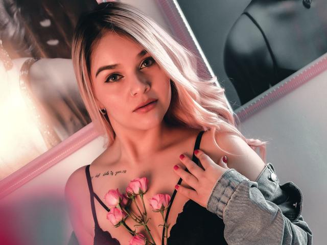 ValerieStone with pink roses, black lace top, and tattoo on chest, wearing gray jacket, looking at camera, blonde hair, pink nails, holding roses, soft light, slightly parted lips.