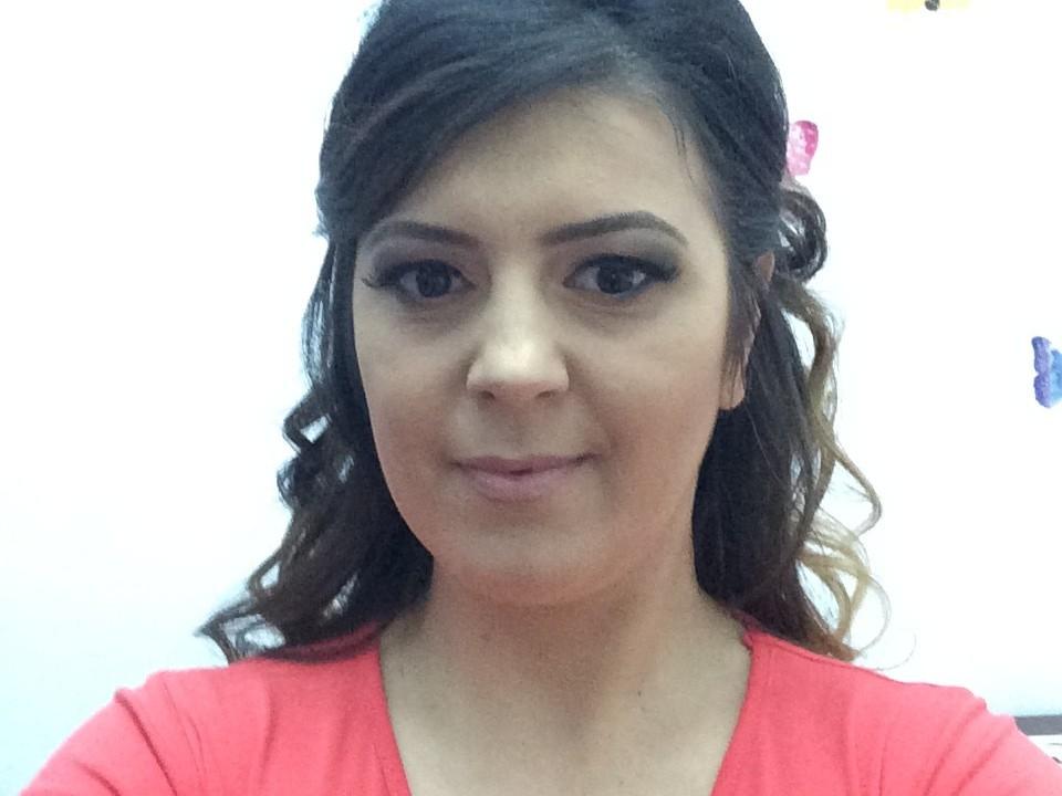 AnneRosse, con i capelli scuri e ondulati, gli occhi marroni e il rossetto rosa, indossa una camicia rossa, ha un leggero sorriso e i suoi seni sono leggermente visibili sotto la camicia..
