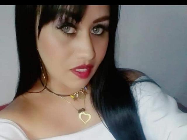 CathyCat, avec ses cheveux noirs, ses yeux verts et son rouge à lèvres rouge, portant un haut blanc, un collier en or avec des pendentifs en forme de cœur et d'étoile, et des boucles d'oreilles créoles, regardant l'appareil photo..