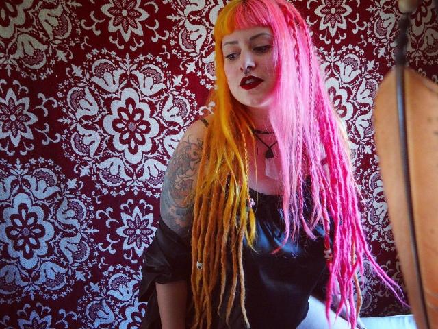SuperSuperHot mit leuchtend rosa und orangefarbenen Dreadlocks, einem schwarzen Lederoberteil, einem tätowierten Arm und dunklem Lippenstift, steht vor einer roten Wand mit floralem Muster und blickt leicht nach unten..