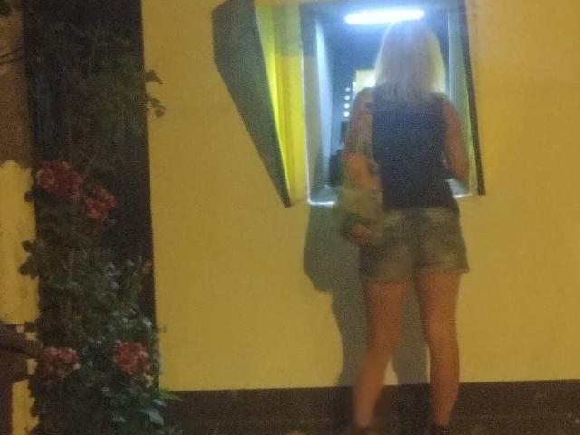 SexyCoco with blonde hair, black sleeveless top, and gray shorts, stands in front of a window, holding a small dog, with yellow curtains and floral decoration.
