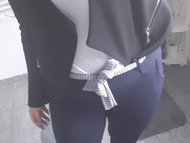 As nádegas de Lynne estão amarradas com um tecido quadriculado preto e branco, ela veste calças escuras e um casaco preto com uma camisa branca. Suas mãos estão ao lado do corpo..