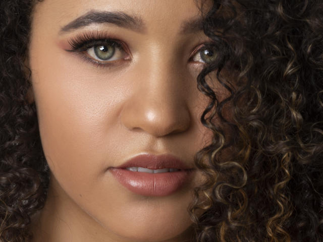 AlyssaBelle's face with curly brown hair, green eyes, and full pink lips, showing a neutral expression. Her skin is smooth with a slight tan, and she's looking forward.