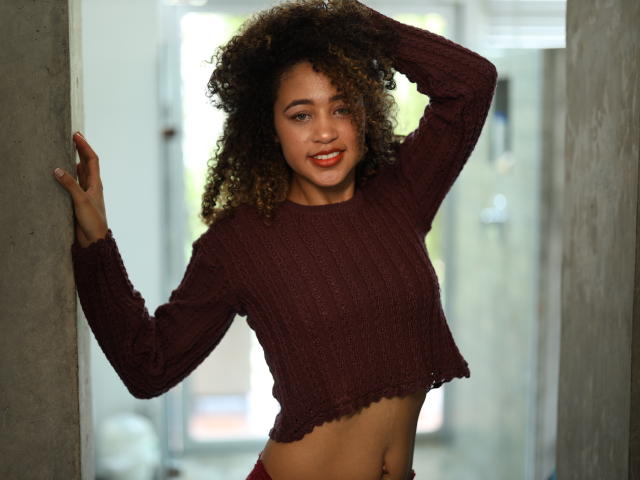 AlyssaBelle mit lockigem, braunem Haar, trägt ein burgunderfarbenes Strick-Crop-Top, das ihren Bauch freilegt, und steht in einem Türrahmen, mit einer Hand auf dem Kopf, lächelnd und mit rotem Lippenstift..