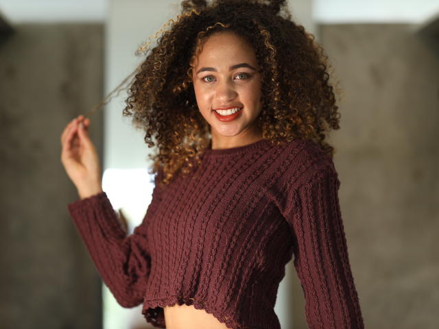 AlyssaBelle, com cabelos castanhos cacheados, sorrindo, vestindo uma blusa cropped de malha marrom, segurando um fio de cabelo, mostrando um rosto fofo e atraente e seus pequenos seios firmes..