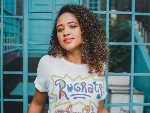 AlyssaBelle tem cabelos castanhos cacheados, pele clara e um sorriso adorável. Ela está vestindo uma camiseta branca de "Rugrats", com pequenos desenhos nela, e está em frente a barras azuis..