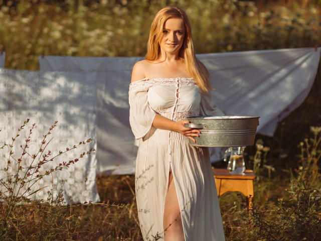 OhMollyHoney, com cabelos loiros longos, vestindo um vestido branco com um ombro descoberto, segurando um balde de metal, em pé em um campo ensolarado, mostrando os seios e uma perna através de uma fenda no vestido..