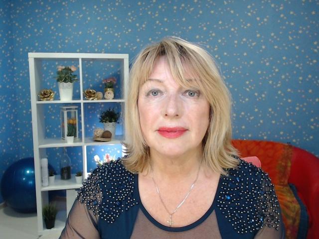 SamanthaLala with blonde hair, blue eyes, and red lipstick, wearing a sheer black top with sparkly shoulders, standing in front of a white shelf with potted plants.