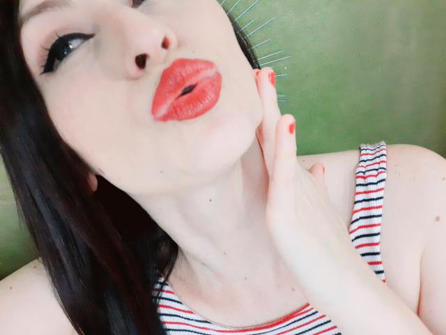 AliceRossi's face, pale skin, red lipstick, black eyeliner, striped tank top, hand touching neck, dark brown hair, green background, red nail polish, pouty lips, slightly tilted head.