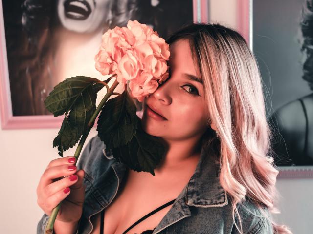 ValerieStone holding pink roses, wearing a denim jacket, black strappy top, pink nails, blonde hair, smiling, green eyes, slightly wavy hair, in a softly lit room.