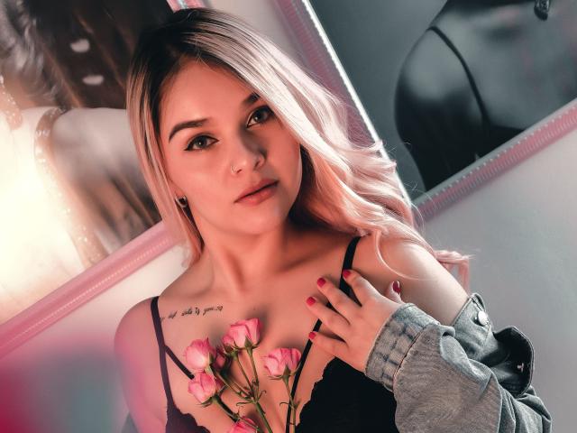 ValerieStone with pink roses on her tits, blonde hair, red nails, black bra, tattoo, light makeup, looking at camera, gray jacket, pink and red light, soft expression.