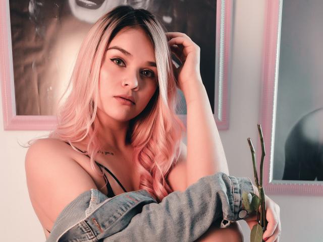 ValerieStone with pink hair, blue denim jacket, black bra, holding a green plant, hand in hair, looking at camera, soft light, slight smile, small tattoo on chest.