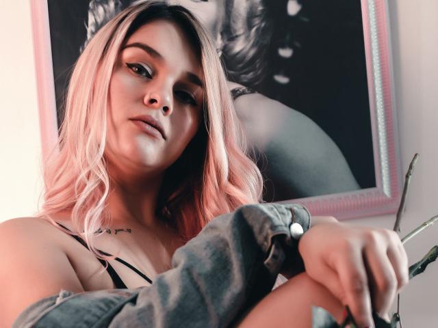 ValerieStone with pink hair, wearing a denim shirt, holding a plant, looking down with a slight smile, black-and-white portrait in the background, tattoo on her chest.