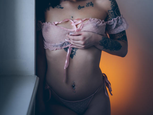 KassiKassou mit Tattoos, trägt rosafarbene, gepunktete Dessous, hält ihre kleinen Brüste, zeigt ihren gestochenen Bauchnabel und ihre seitlich gebundenen Slips, in weichem, gedämpftem Licht..