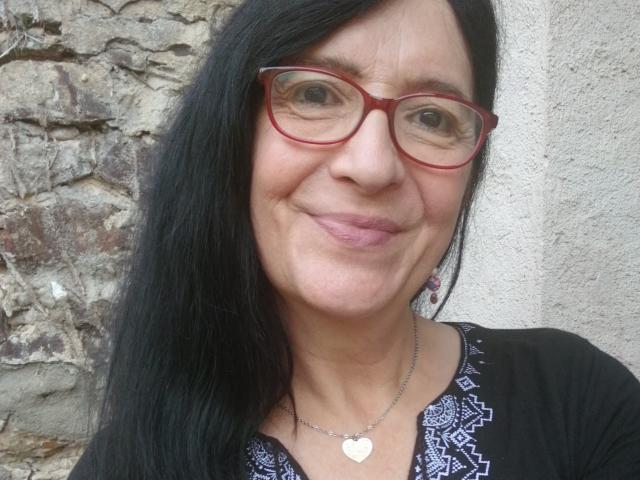 Emilianne, com óculos vermelhos, cabelo preto, sorrindo, lábios rosados e cheios, colar com formato de coração, blusa preta com estampa, pele levemente enrugada, em pé em frente a uma parede de pedra áspera..