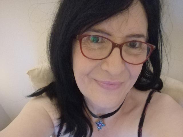 Emilianne, avec ses cheveux noirs, ses lunettes rouges, sa peau claire, un léger sourire, portant un collier noir avec un cœur bleu, et un bandeau laissant entrevoir ses seins nus..