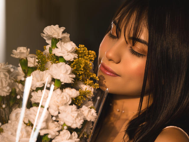 VanityValos mit geschlossenen Augen, die einen Strauß aus weißen Blumen und gelben Knospen hält, wobei ihr dunkles Haar und ihre glänzenden, rosafarbenen Lippen im weichen Licht einen Kontrast bilden..