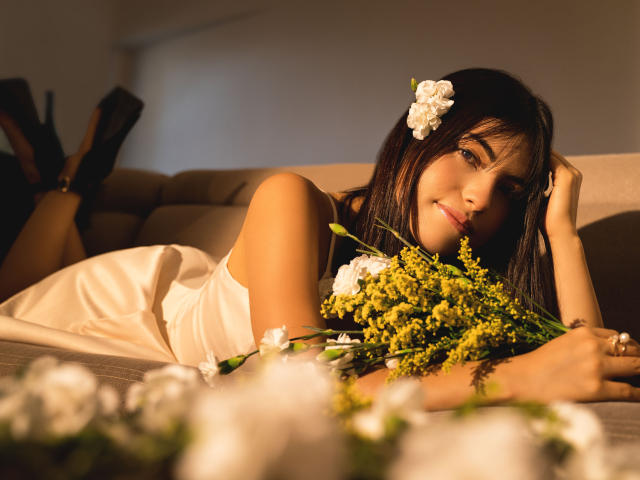 VanityValos, liegend auf einer Couch, trägt ein weißes Kleid, hält gelbe Blumen, hat eine weiße Blume in ihrem dunklen Haar, trägt schwarze High Heels, lächelt, warmes Licht..