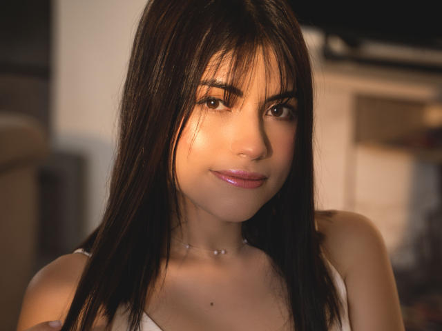 VanityValos, com cabelo castanho escuro, franja e uma camiseta branca, exibe um sorriso adorável, lábios rosados e uma pequena pinta no peito. Ela está usando um colar delicado..
